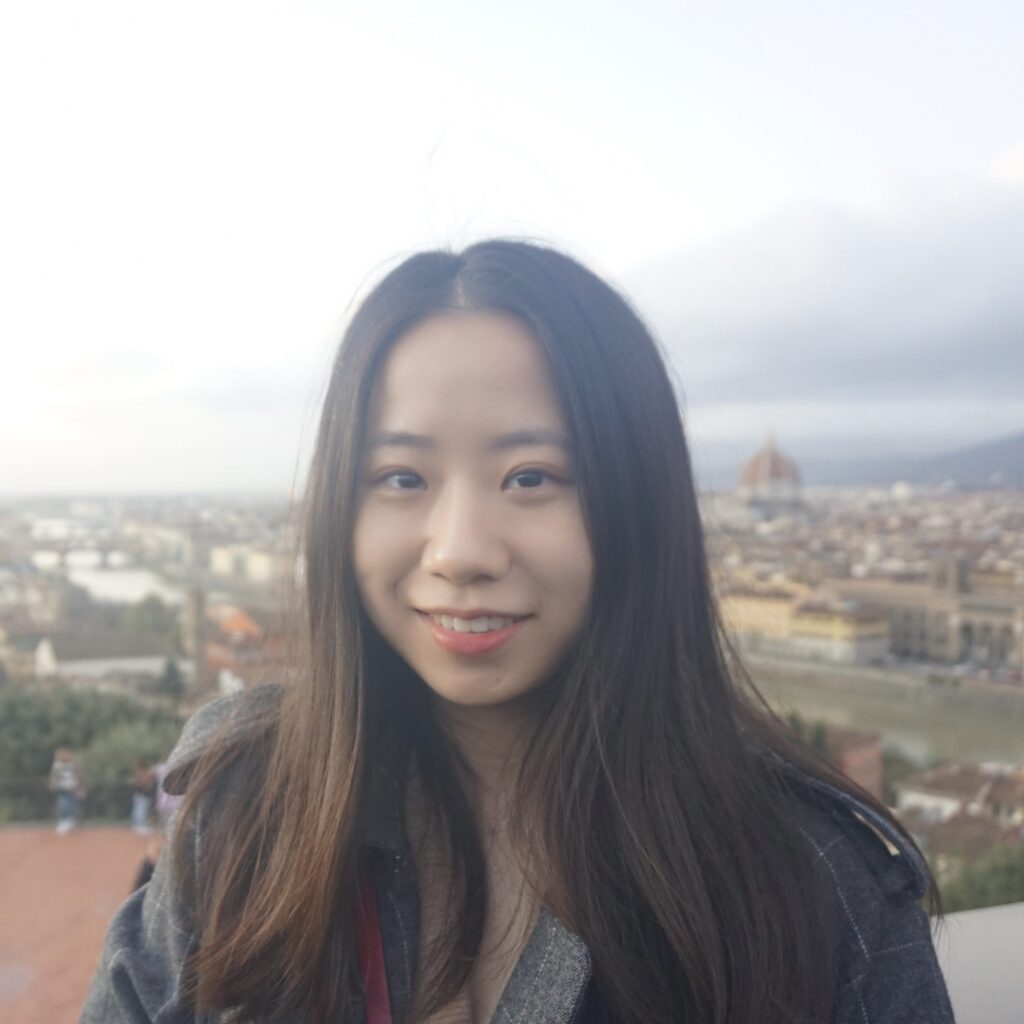 A young woman with long dark hair smiles in front of a cityscape with historic buildings and a dome under a cloudy sky.