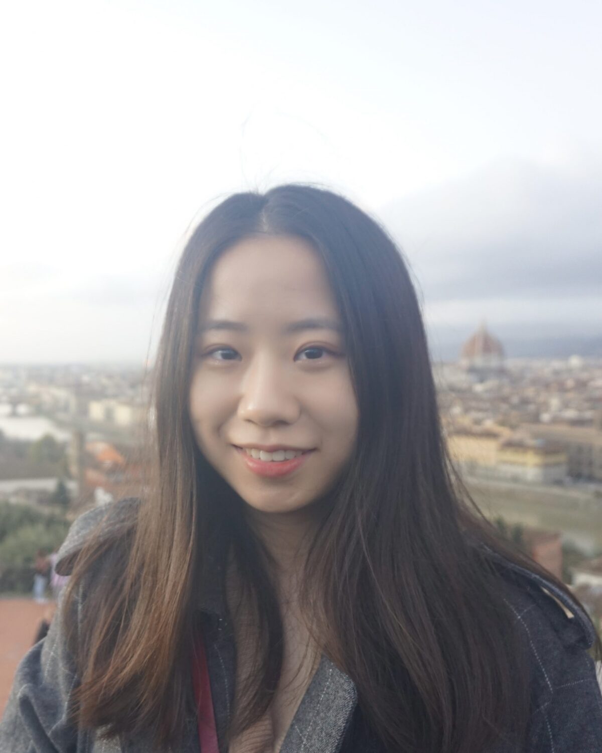 A young woman with long dark hair smiles in front of a cityscape with historic buildings and a dome under a cloudy sky.