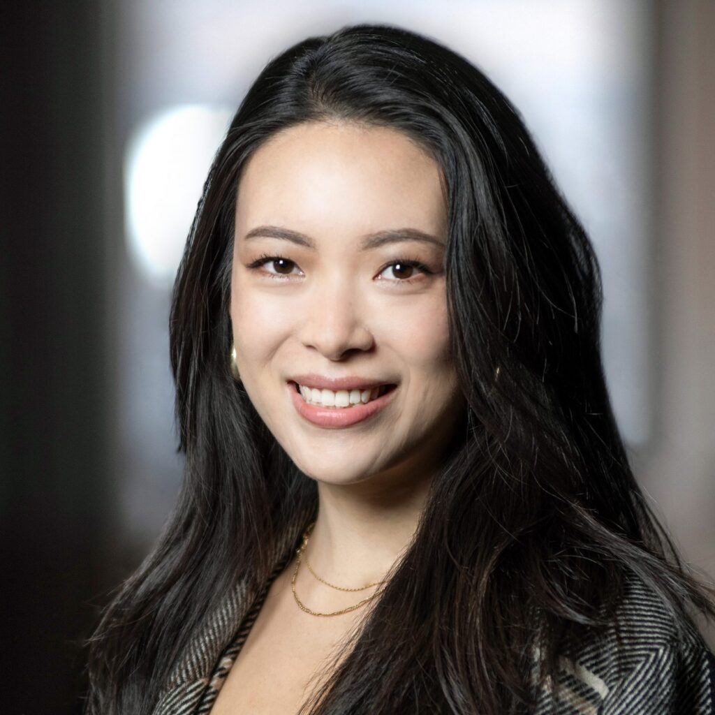 A woman with long dark hair smiles at the camera, wearing a patterned top and gold necklaces, with a softly blurred background.