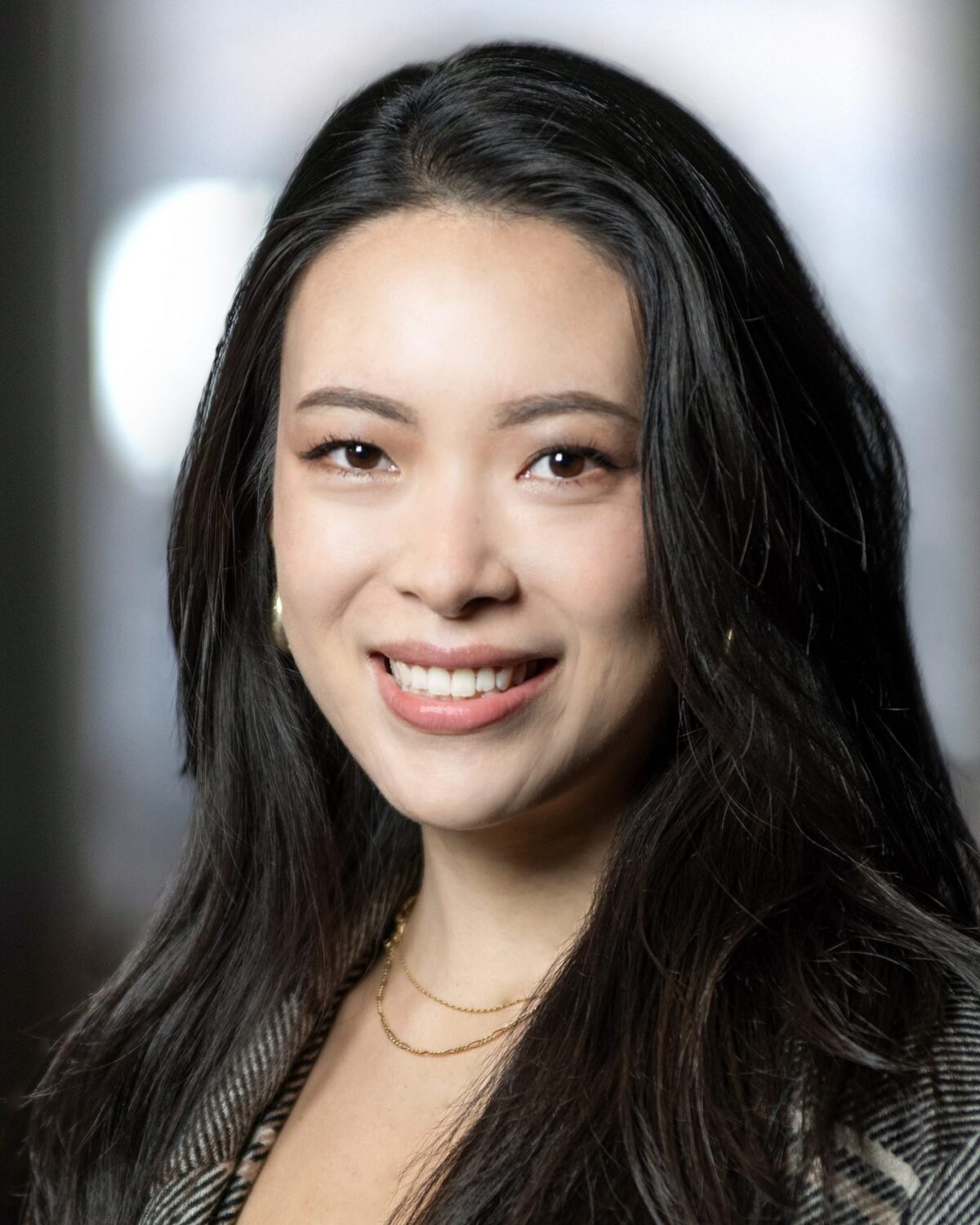 A woman with long dark hair smiles at the camera, wearing a patterned top and gold necklaces, with a softly blurred background.
