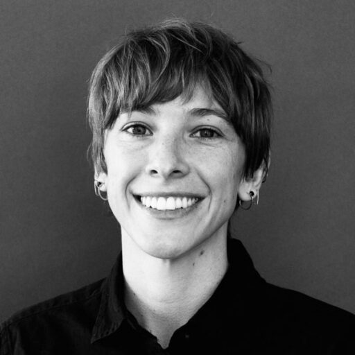 Smiling person with short, light brown hair wearing a dark collared shirt and small hoop earrings, standing in front of a plain dark background.