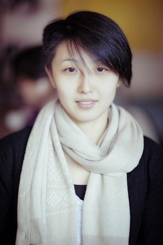 A person with short black hair, wearing a light beige scarf and a black sweater, looks at the camera with a slight smile. The background is softly blurred.