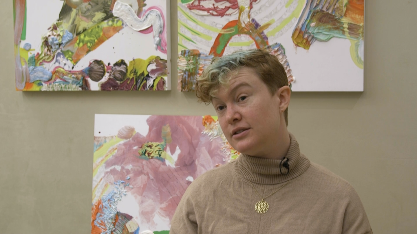A person with short, curly hair and a beige turtleneck sits in front of colorful abstract paintings, looking slightly to the side.