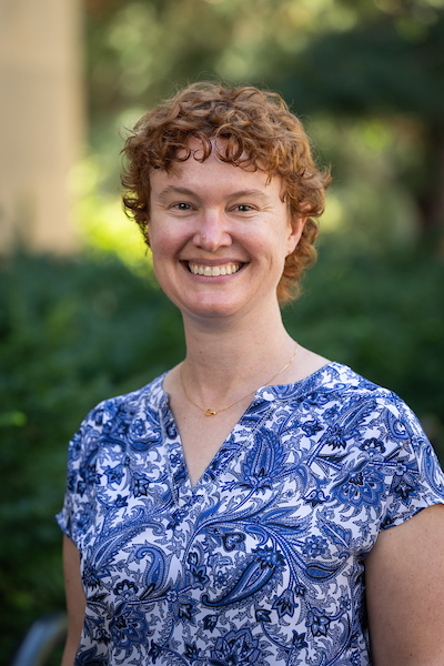 A person with short curly red hair, wearing a blue and white paisley-patterned shirt, smiles at the camera. Green foliage and soft sunlight fill the background.