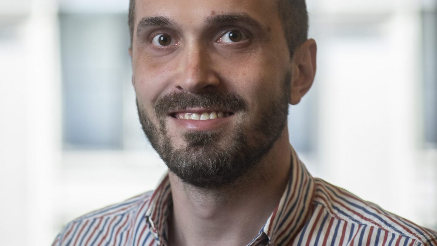 A man with a beard and short hair, wearing a striped button-up shirt, smiles at the camera. The background is blurred with large bright windows.