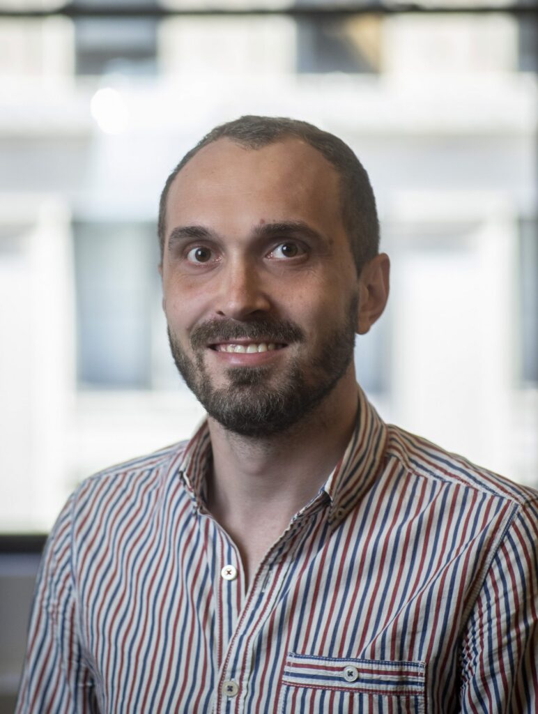 A man with a beard and short hair, wearing a striped button-up shirt, smiles at the camera. The background is blurred with large bright windows.