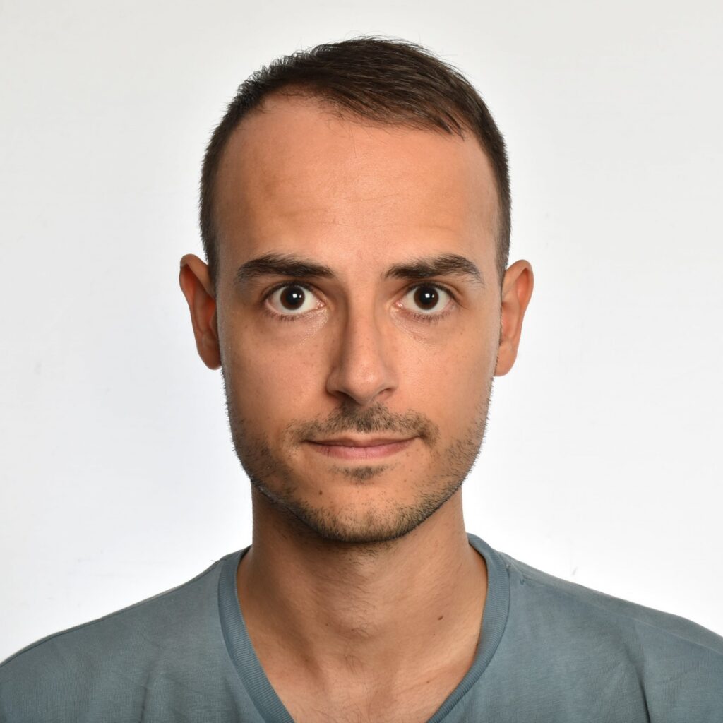 A man with short brown hair and a trimmed beard looks directly at the camera. He is wearing a light blue T-shirt and stands against a plain white background.
