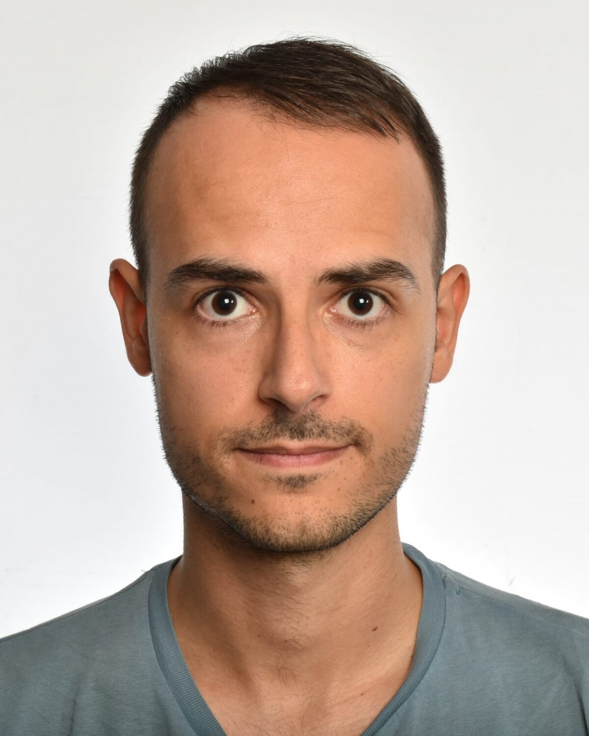 A man with short brown hair and a trimmed beard looks directly at the camera. He is wearing a light blue T-shirt and stands against a plain white background.