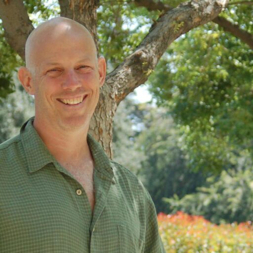 A smiling bald man in a green shirt stands outdoors in front of leafy trees and bushes on a sunny day.