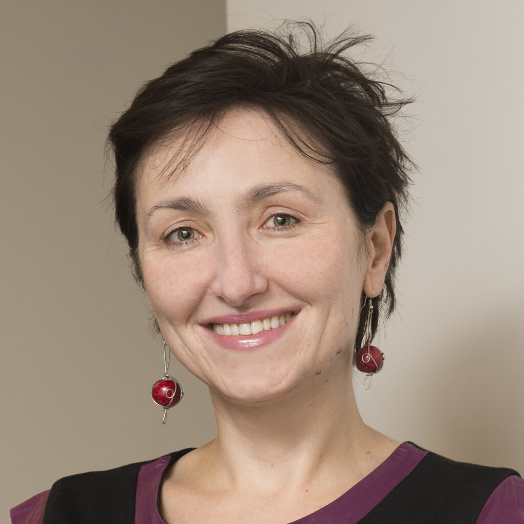 A woman with short brown hair smiles at the camera. She is wearing red bead earrings and a black top with a purple neckline, standing in front of a neutral-colored wall.