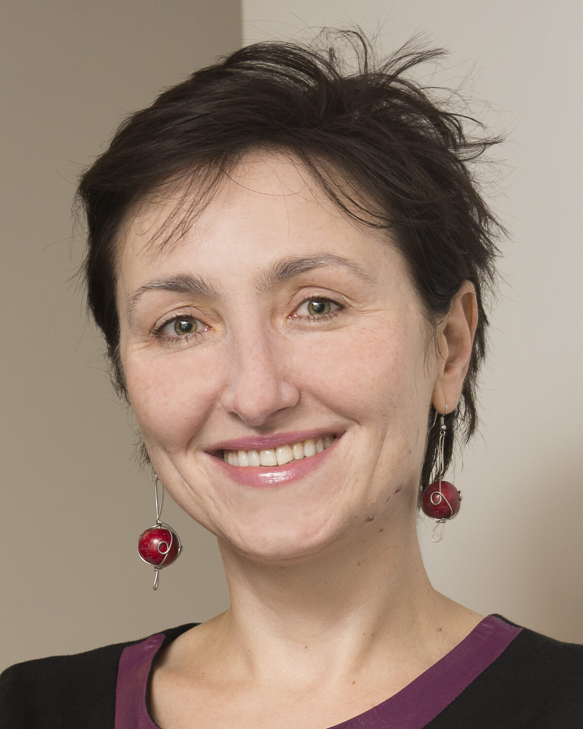 A woman with short brown hair smiles at the camera. She is wearing red bead earrings and a black top with a purple neckline, standing in front of a neutral-colored wall.