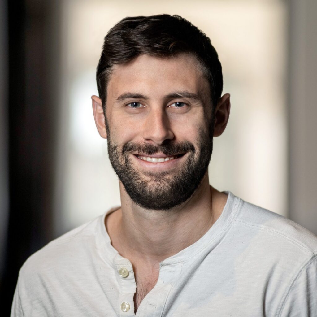 A man with short brown hair and a beard, wearing a light-colored henley shirt, smiles at the camera. The background is softly blurred.