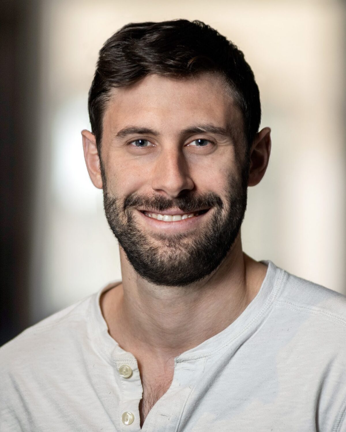 A man with short brown hair and a beard, wearing a light-colored henley shirt, smiles at the camera. The background is softly blurred.