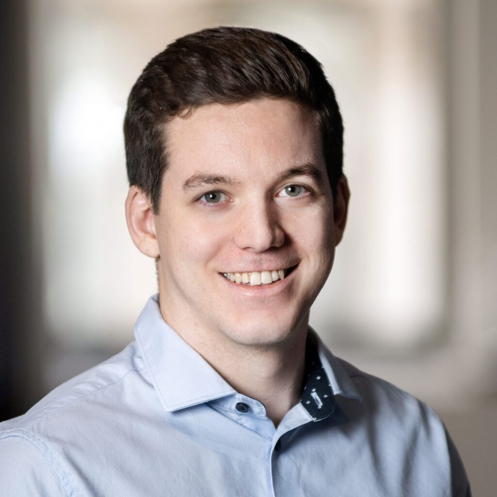 A young man with short brown hair smiles, wearing a light blue button-up shirt in a softly lit indoor setting with a blurred background.