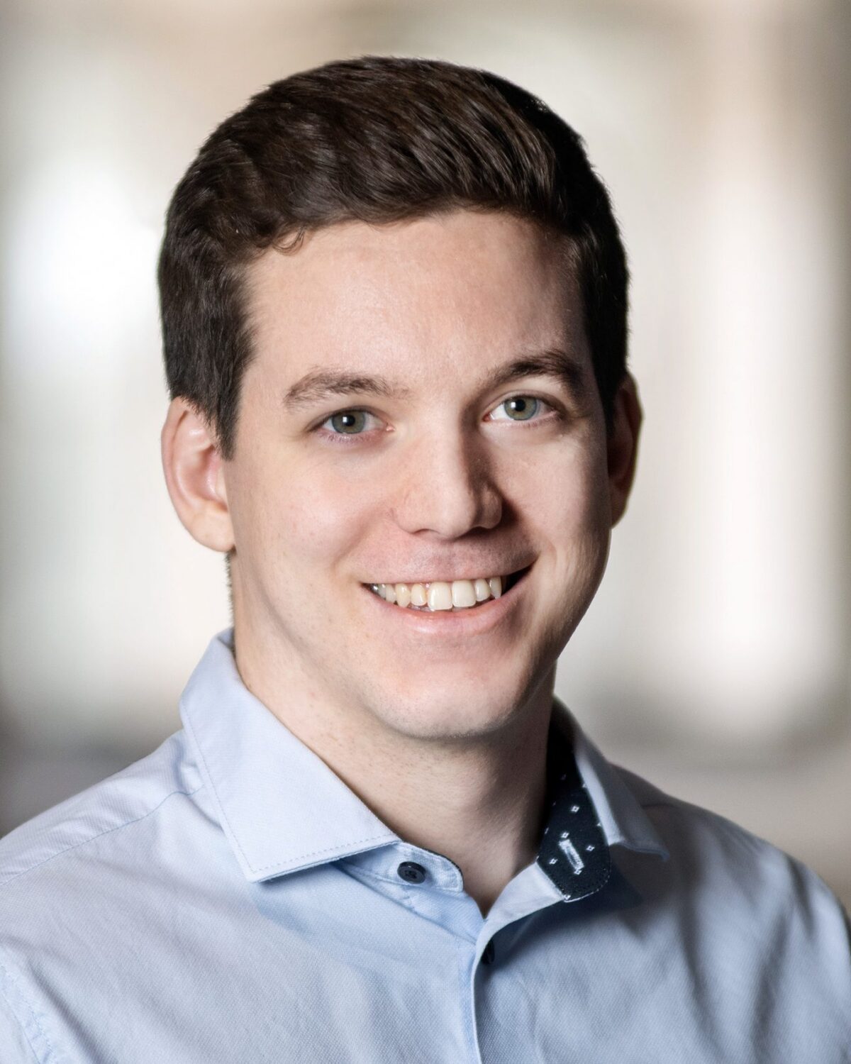 A young man with short brown hair smiles, wearing a light blue button-up shirt in a softly lit indoor setting with a blurred background.