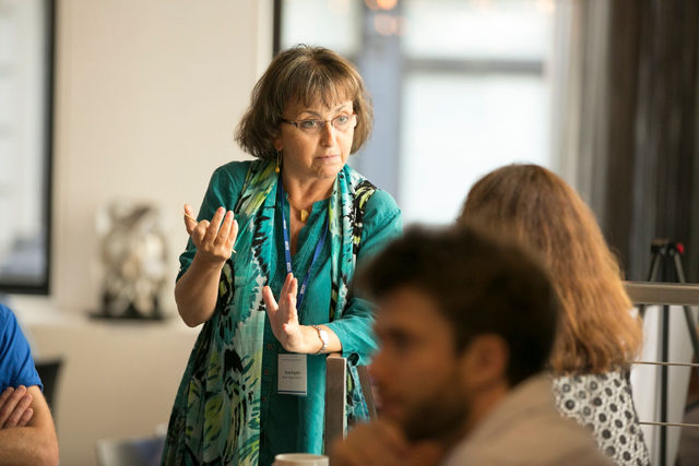 A woman wearing glasses and a blue patterned scarf gestures while speaking to two people in a brightly lit indoor setting. Other people are visible in the foreground.