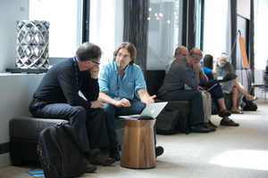 Two people sit on a bench having a conversation, one with a laptop open. Other pairs of people are talking in the background along the same bench in a well-lit modern room.