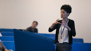 A person with short hair presents in front of a class, gesturing with hands near a podium. Several people sit in blue seats, listening attentively.