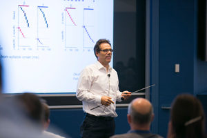 A man in a white shirt gives a presentation in a conference room, holding a pointer. Graphs are displayed on a large screen behind him. Several audience members are visible from behind.