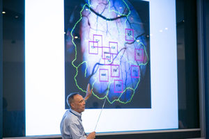 A person points at a large image of a brain on a screen, which is outlined in green with several red squares marking different regions.