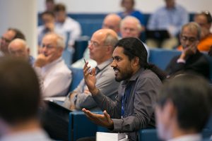 A man with long hair speaks and gestures while seated among a group of people in a conference or lecture setting. Other attendees listen attentively in the background.