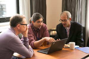 Three men sit around a wooden table, focused on a laptop screen. One man gestures at the laptop while the others listen. A coffee mug, plate, and notepad are on the table.