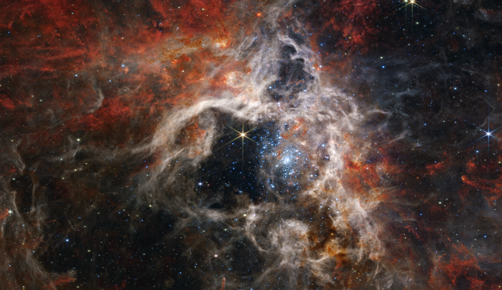 A vibrant star cluster shrouded by red, orange, and gray clouds of cosmic gas and dust in deep space, with numerous stars scattered across the scene.