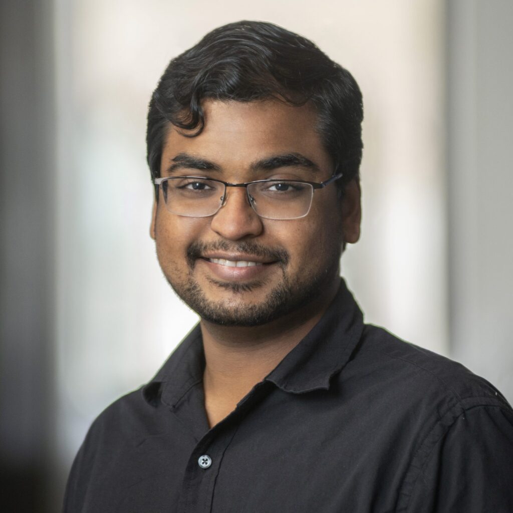 A man with short dark hair, glasses, and a trimmed beard smiles at the camera. He is wearing a black collared shirt and standing in front of a softly blurred indoor background.