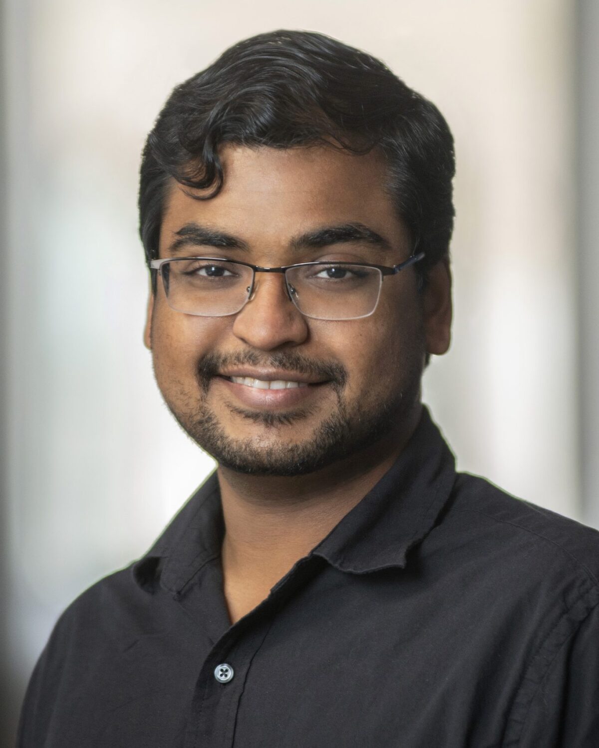 A man with short dark hair, glasses, and a trimmed beard smiles at the camera. He is wearing a black collared shirt and standing in front of a softly blurred indoor background.