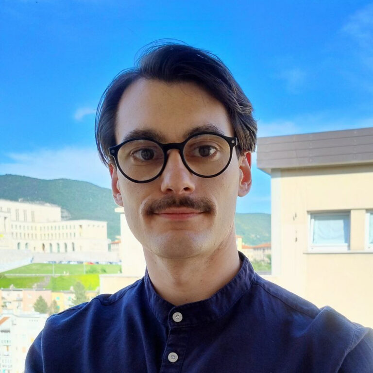 A man with dark hair, glasses, and a mustache stands outside with buildings, greenery, and mountains in the background under a clear blue sky.