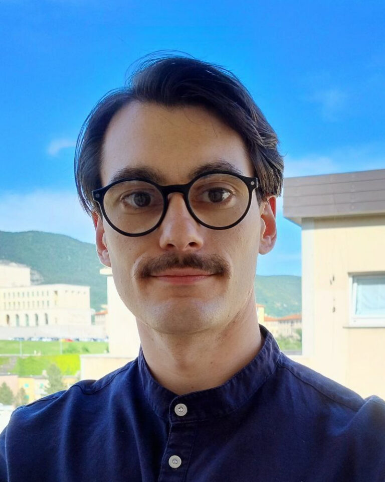 A man with dark hair, glasses, and a mustache stands outside with buildings, greenery, and mountains in the background under a clear blue sky.