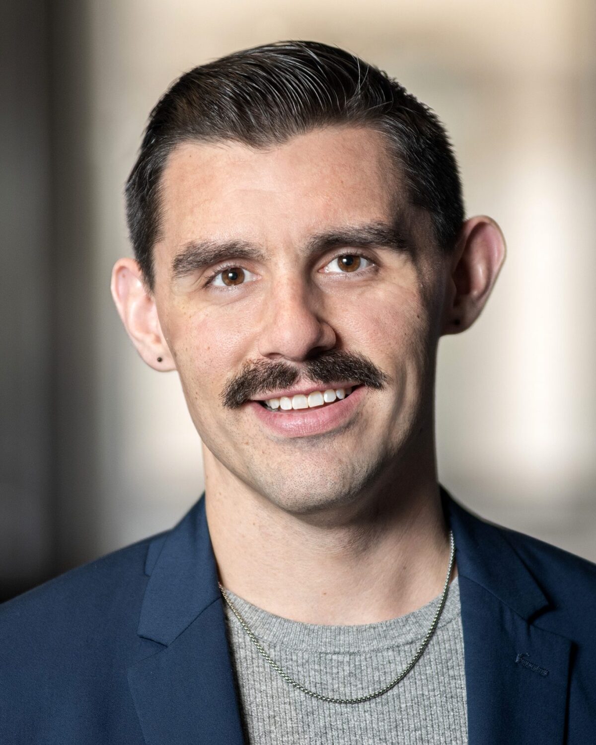 A man with dark hair and a mustache, wearing a blue blazer, gray shirt, and a chain necklace, smiles at the camera with a blurred neutral background.
