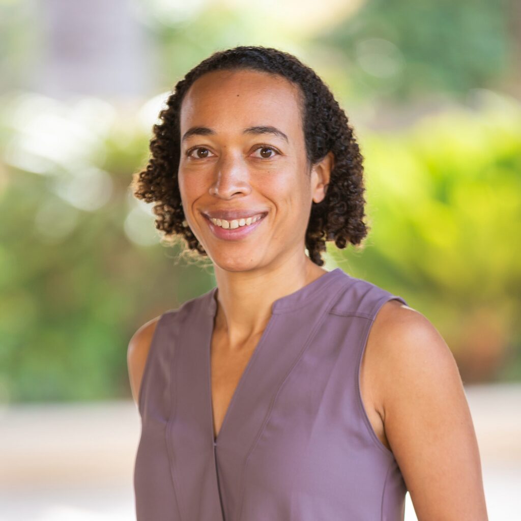 A woman with curly short hair, wearing a sleeveless lavender top, smiles at the camera. The background is softly blurred with green and yellow foliage.