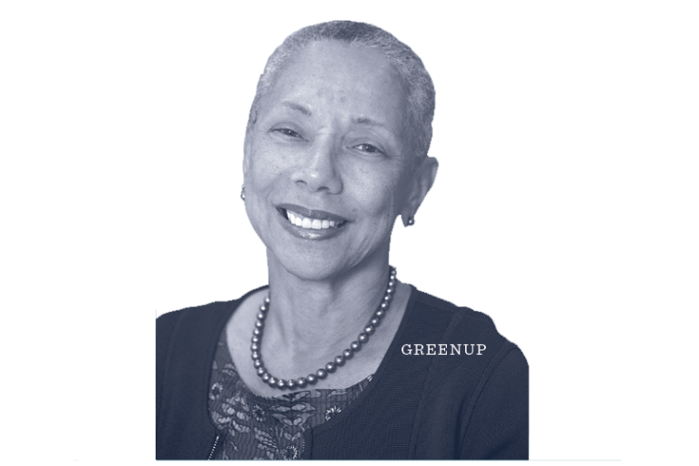 Smiling older woman with short gray hair, wearing a necklace and dark top, against a light background. The word GREENUP appears near her shoulder.