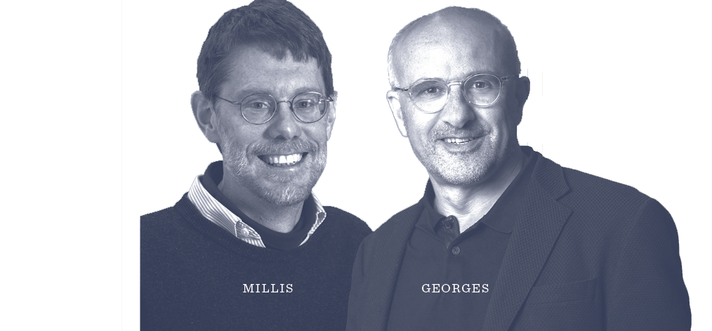 Two men wearing glasses, one in a sweater and shirt labeled Millis, the other in a jacket and shirt labeled Georges, stand next to each other, smiling at the camera against a plain background.