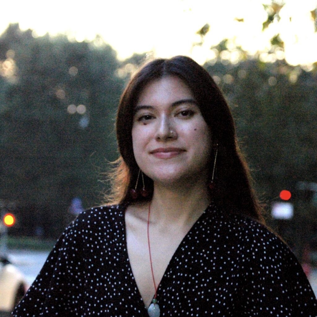 A person with long brown hair, wearing a black and white polka dot top, stands outdoors on a city street at dusk, smiling softly at the camera. Trees and blurred cars are visible in the background.