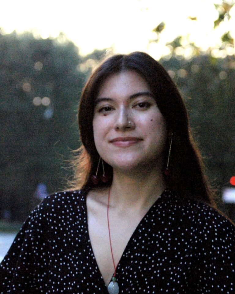 A person with long brown hair, wearing a black and white polka dot top, stands outdoors on a city street at dusk, smiling softly at the camera. Trees and blurred cars are visible in the background.