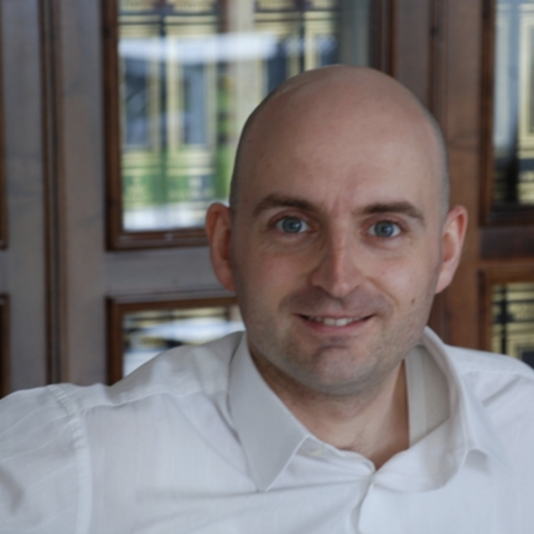 A man with a shaved head, wearing a white shirt, smiles at the camera. Behind him are shelves filled with books in black and gold bindings.