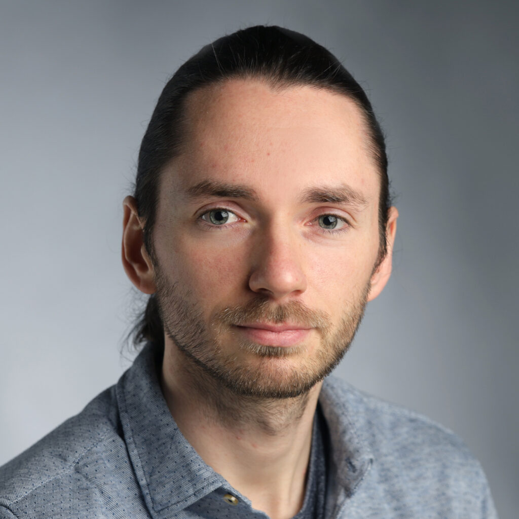 A man with long brown hair tied back, light beard, and blue eyes, wearing a grey collared shirt, posed against a plain, softly lit background.