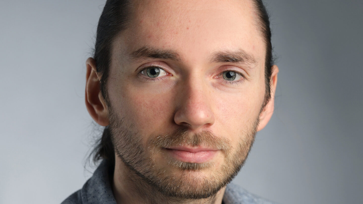 A man with long brown hair tied back, light beard, and blue eyes, wearing a grey collared shirt, posed against a plain, softly lit background.