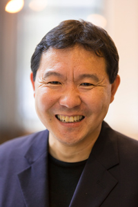 A middle-aged man with short black hair smiles warmly at the camera. He is wearing a dark blazer over a black shirt, and the background is softly blurred.