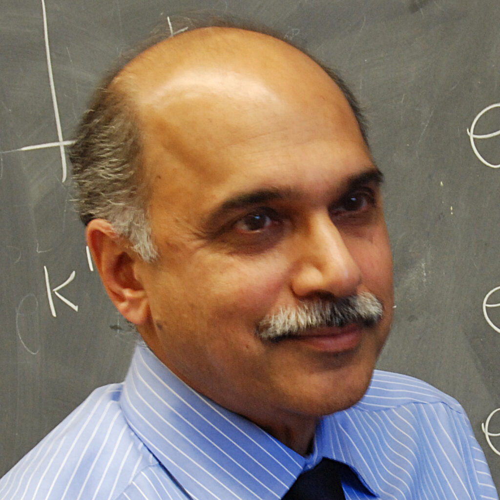 A man with a mustache and short hair wearing a blue striped shirt and black tie stands in front of a chalkboard with writing on it.