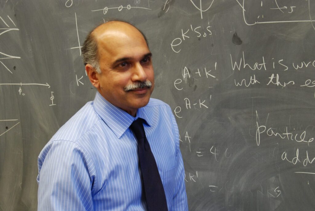 A man in a blue striped shirt and dark tie stands in front of a chalkboard covered with scientific equations and diagrams.
