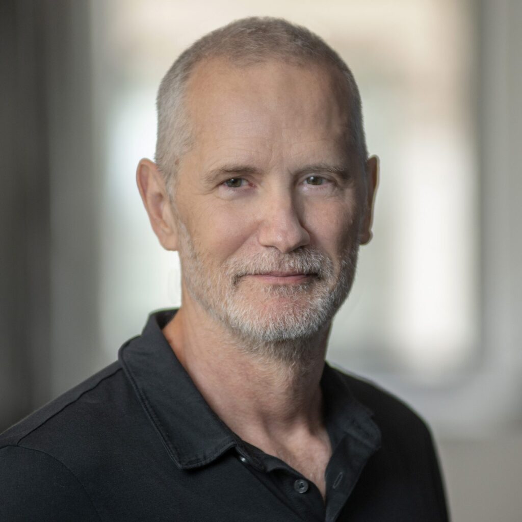 A middle-aged man with short gray hair and a beard, wearing a black collared shirt, stands indoors with a soft, blurred background.