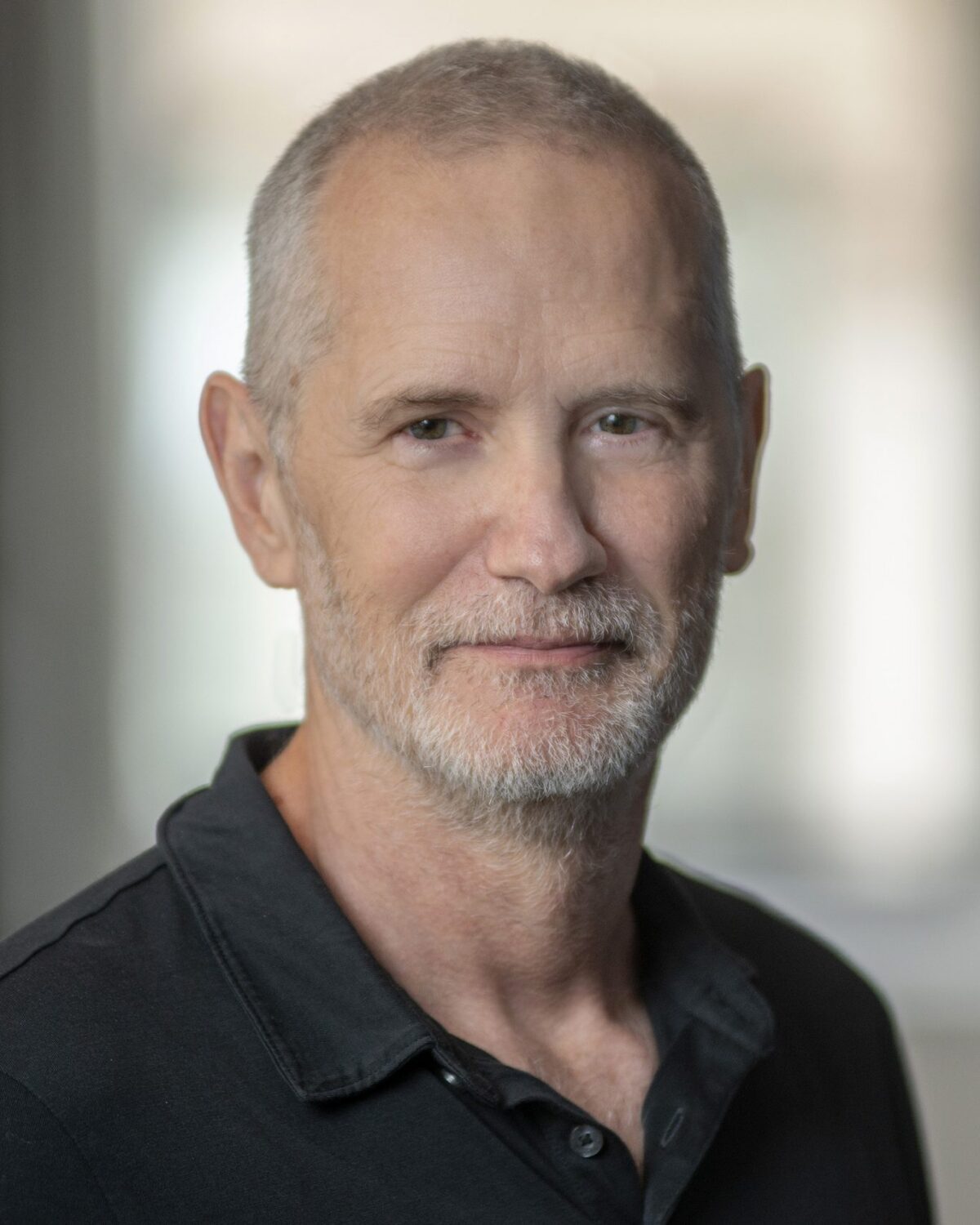 A middle-aged man with short gray hair and a beard, wearing a black collared shirt, stands indoors with a soft, blurred background.