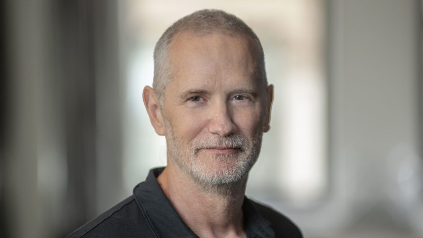 A middle-aged man with short gray hair and a beard, wearing a black collared shirt, stands indoors with a soft, blurred background.