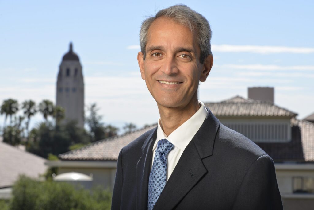 A man in a suit and tie smiles outdoors with tiled rooftops and a tower in the background under a clear sky.