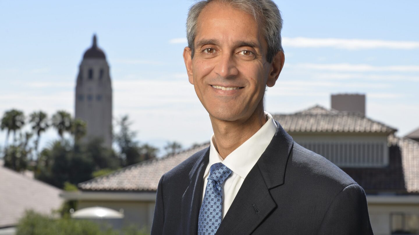 A man in a suit and tie smiles outdoors with tiled rooftops and a tower in the background under a clear sky.