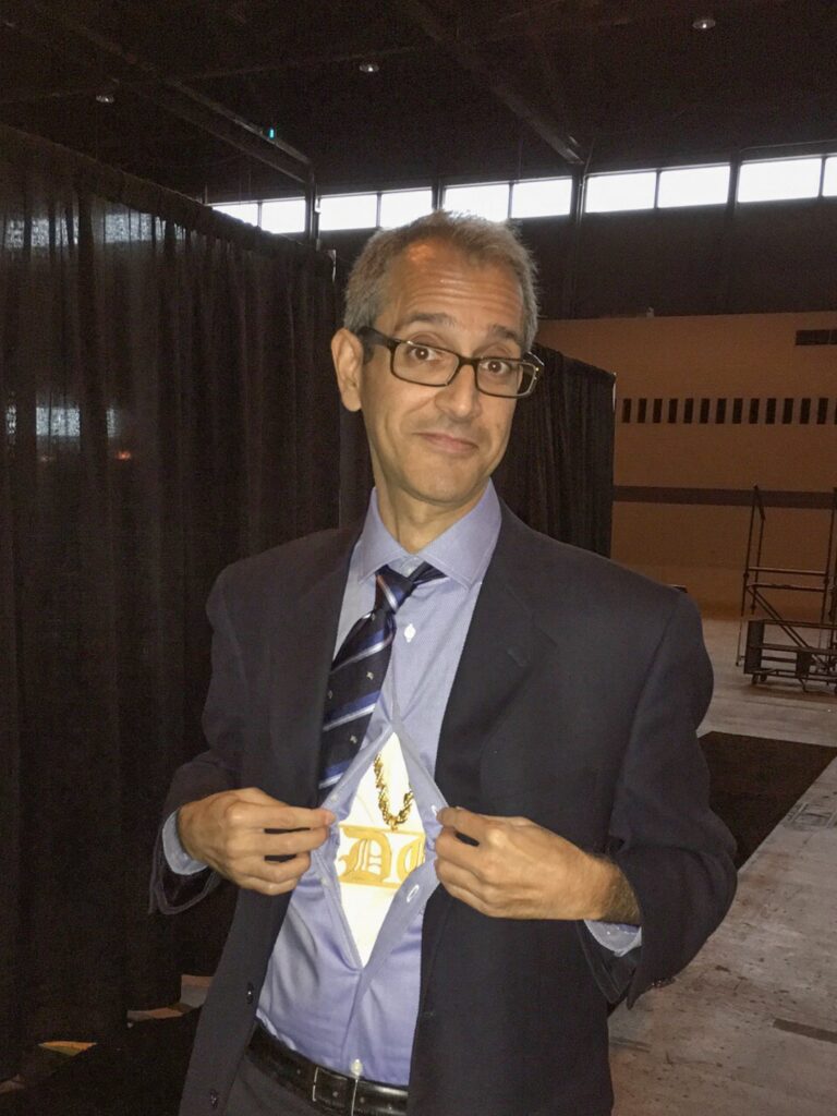 A man in a suit jacket opens his shirt to reveal a superhero emblem underneath, standing indoors with black curtains and a stage behind him.