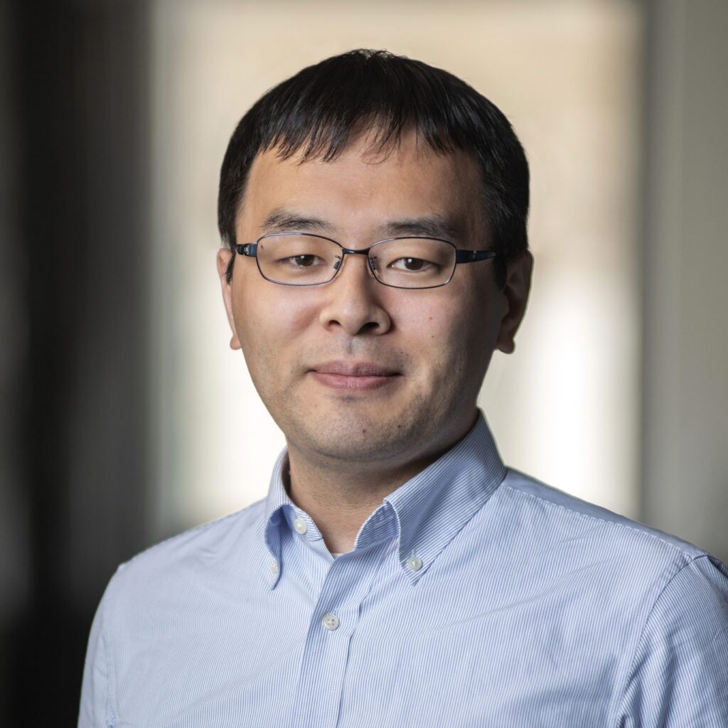 A man with short dark hair and glasses, wearing a light blue button-up shirt, stands indoors with a neutral expression. The background is softly blurred.
